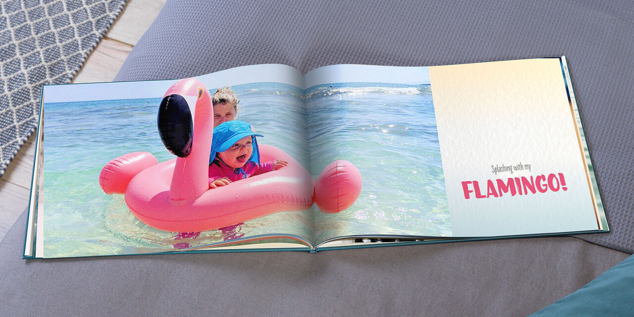 An open CEWE PHOTOBOOK shows Hannah on an inflatable flamingo in the pool. On the right side of the book is written "Splashing with my FLAMINGO!".