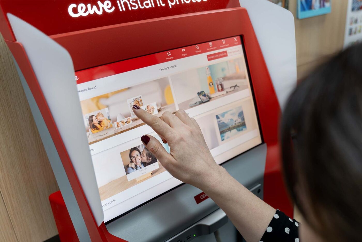 A hand taps the screen of a CEWE instant photo kiosk.