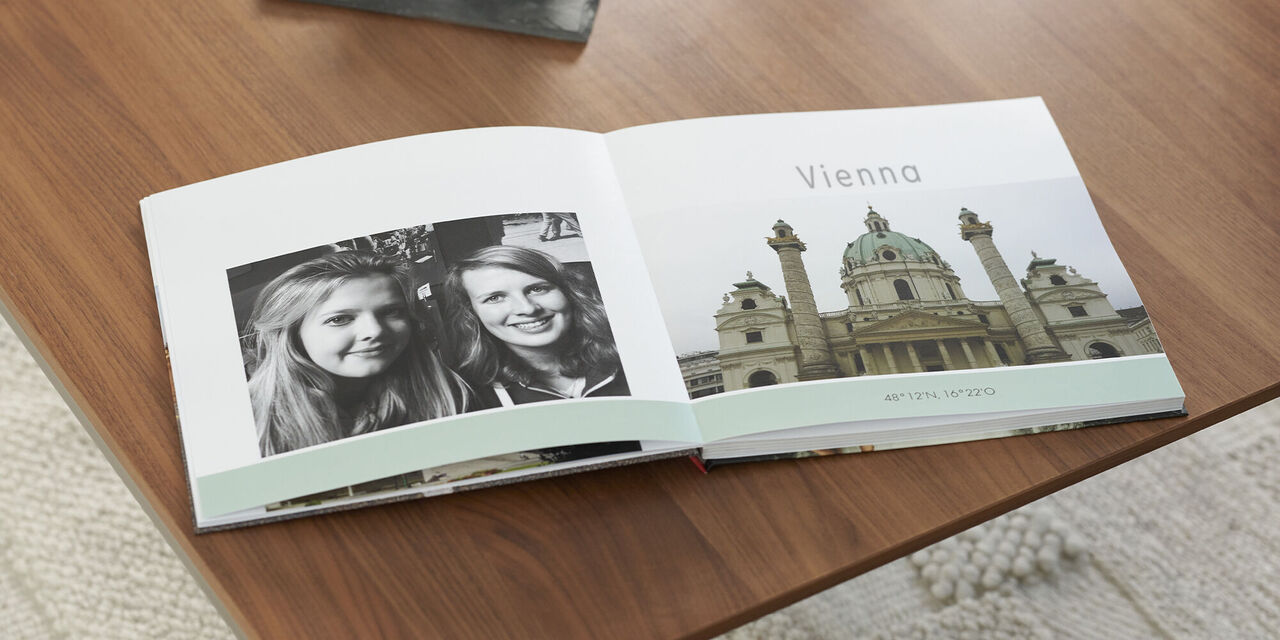 On a wooden table lies an open photo book with a black and white picture of two women and a picture of a church as well as the headline Vienna. Above the photo book is a dark tray with a candle and shell. Under the table you can see a bright carpet.