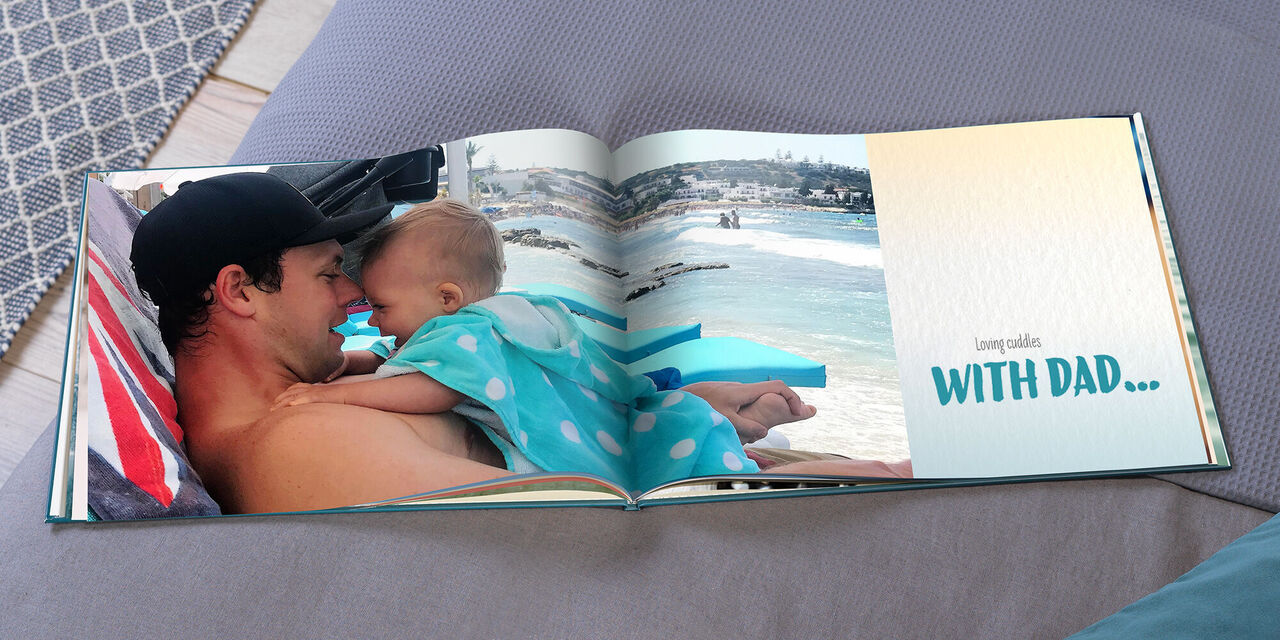 An open CEWE PHOTOBOOK shows Evan on the beach, his daughter Hannah lying on him. On the right side of the book is written "Loving cuddling WITH DAD..."