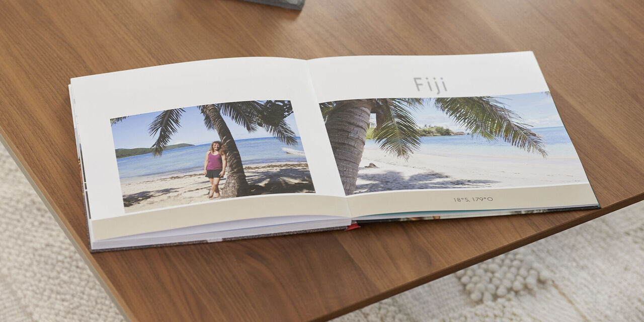 On a wooden table lies an open photo book with two beach pictures and the headline Fiji. Above the photo book is a dark tray with a candle and shell. Under the table you can see a bright carpet.